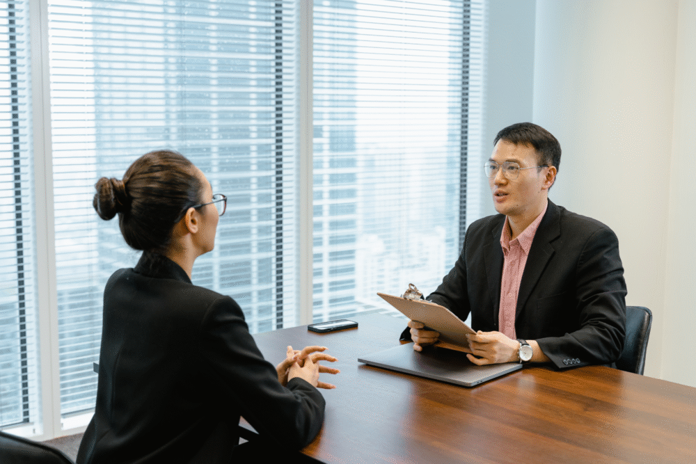 Modern office, interview at desk, large windows frame cityscape, daytime natural light, editorial travel photo, two people.