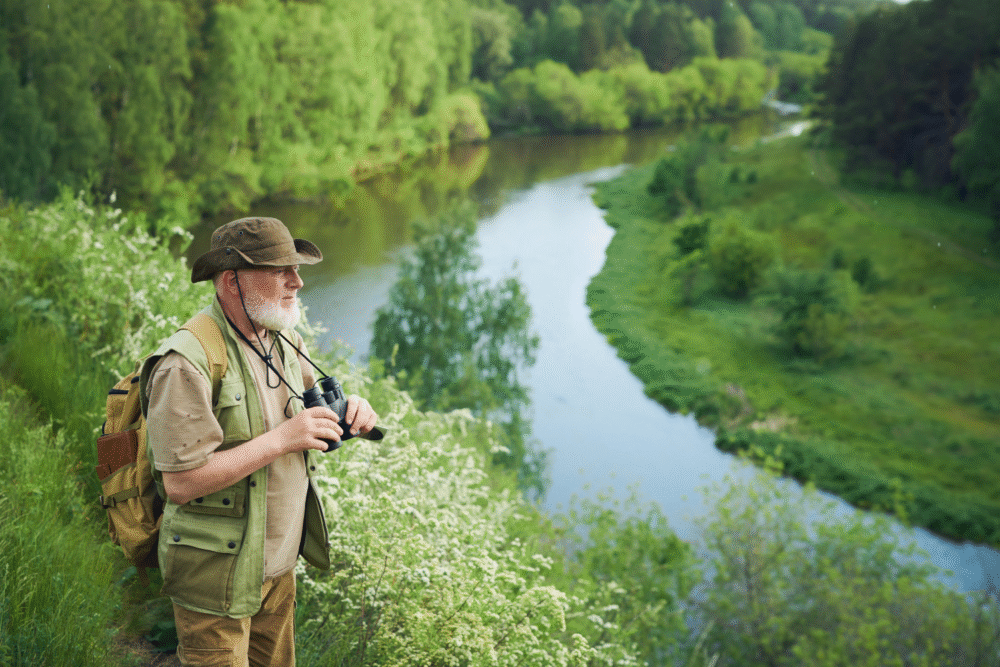 Hillside by winding river, older man with binoculars foreground, verdant midday, editorial travel photo, one person.