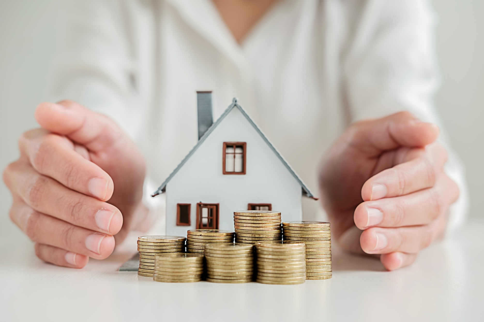 Tabletop, model house and gold coins, close-up encircled by hands, daylight, documentary style, no people.