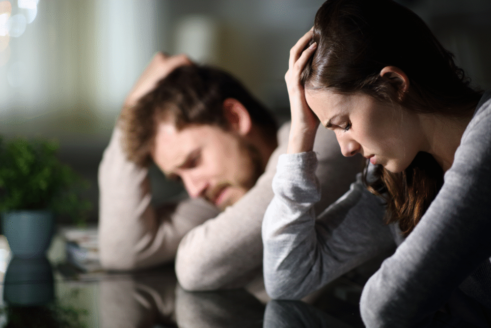Dim café, stressed couple with heads bowed, close composition, afternoon low light, documentary style, two people.