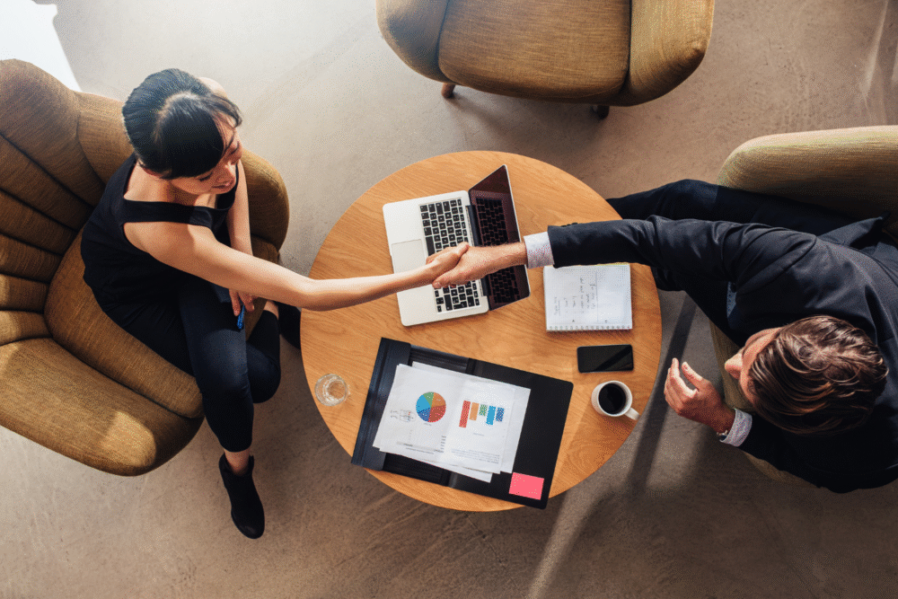 Office meeting room, handshake across round table, overhead view, natural light, documentary style, two professionals.