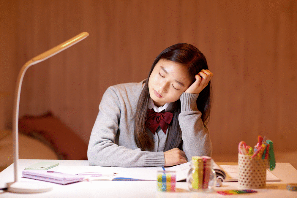 School desk, student resting head, books and stationery scattered, warm lamplight evening, documentary style, one person.