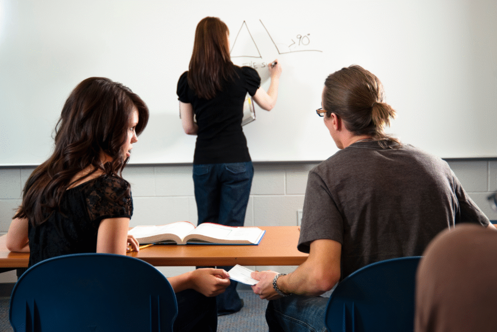 Classroom, student at whiteboard drawing triangle, peers at desk with open book, natural daylight, documentary style, people.
