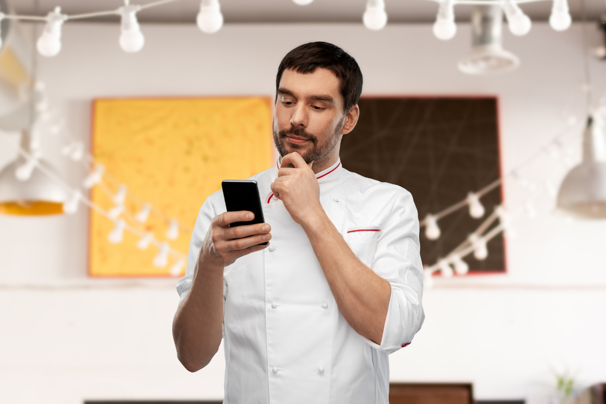 Indoor restaurant, chef in white uniform studies smartphone, mid-shot, bright lights, editorial travel photo, one person.