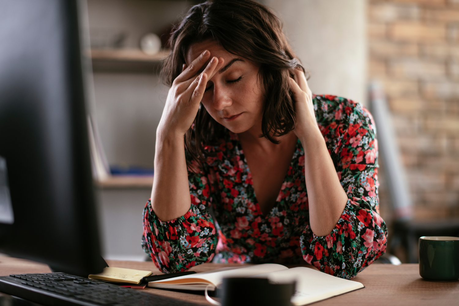 Office desk, stressed woman clutching head, side view with notebook and monitor, daylight, documentary style, people present.