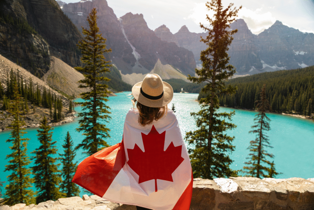 Turquoise alpine lake, Canadian flag draped figure center, wide shot, midday sun, editorial travel photo, single person.