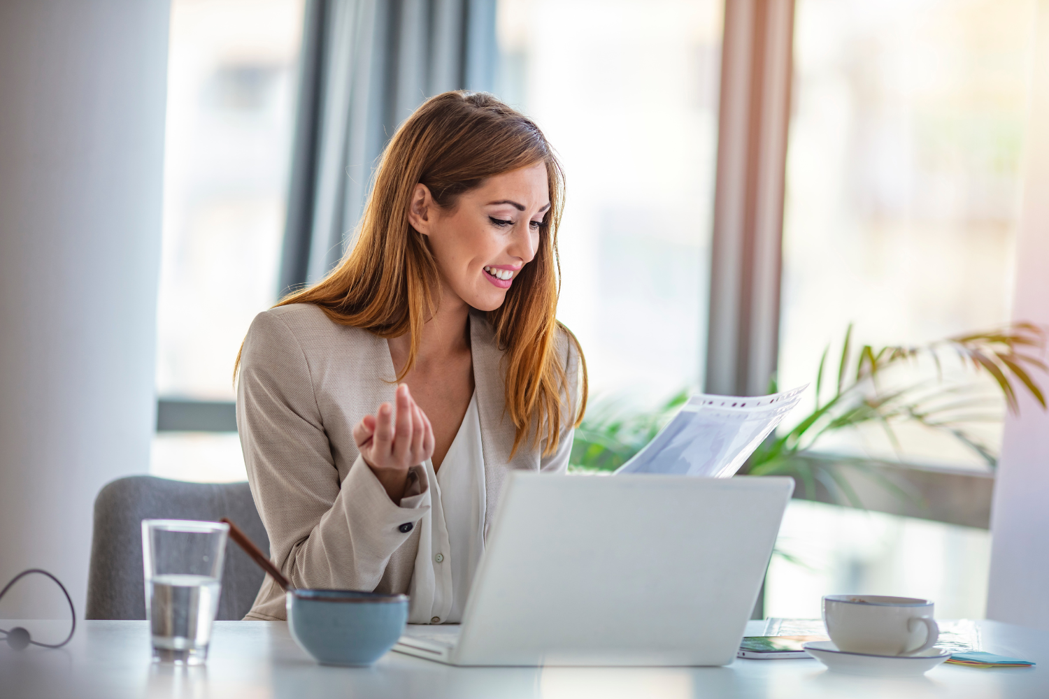 Modern office, woman in beige blazer with laptop, desk setup, daylight bright, documentary style photo, with people.