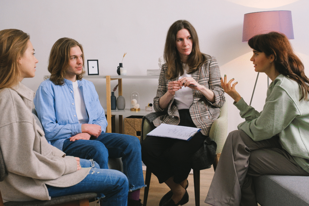 Counseling room, clipboard in woman’s hand, four-person seated circle, daytime indoor light, documentary style, people.
