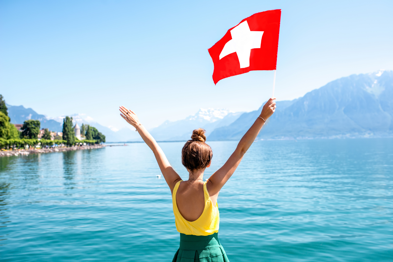 Swiss lakeshore, woman with Swiss flag, wide view over snow peaks and village, midday sun, editorial travel photo, one person.
