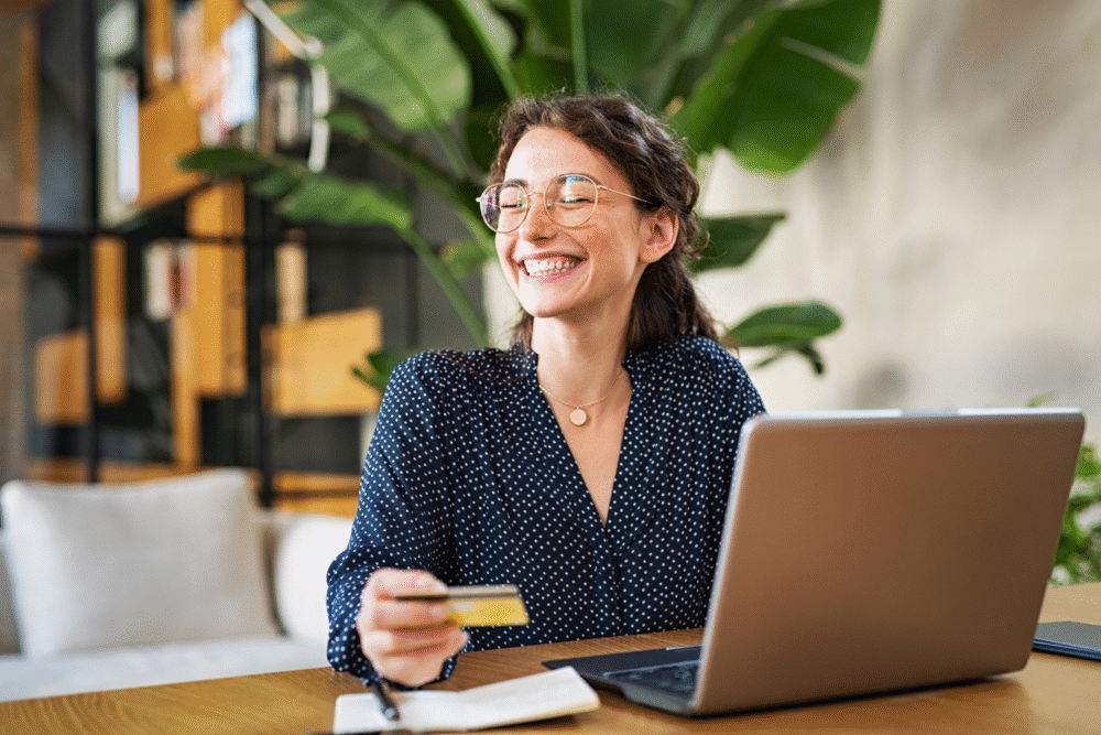 Modern office, woman with glasses at laptop, credit card raised, daylight glow, editorial travel photo, one person.