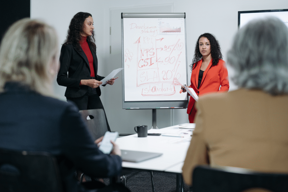 Meeting room, flip chart with notes and percentages, foreground listeners, morning light, documentary style, with people.