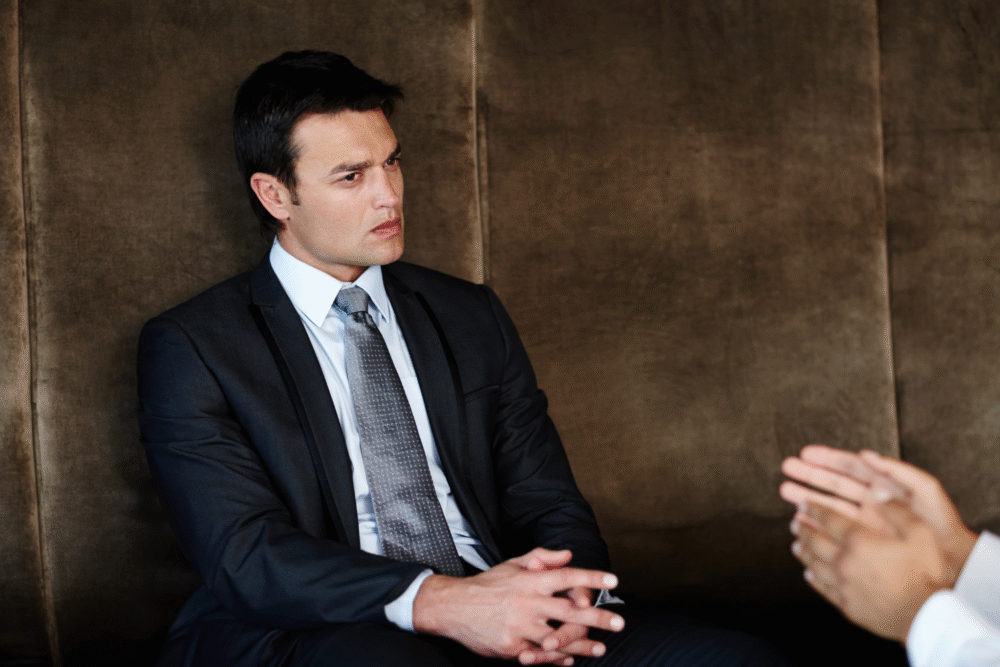 Office boardroom, man in dark suit center frame, hands clasped, midday natural light, documentary style, two people present.