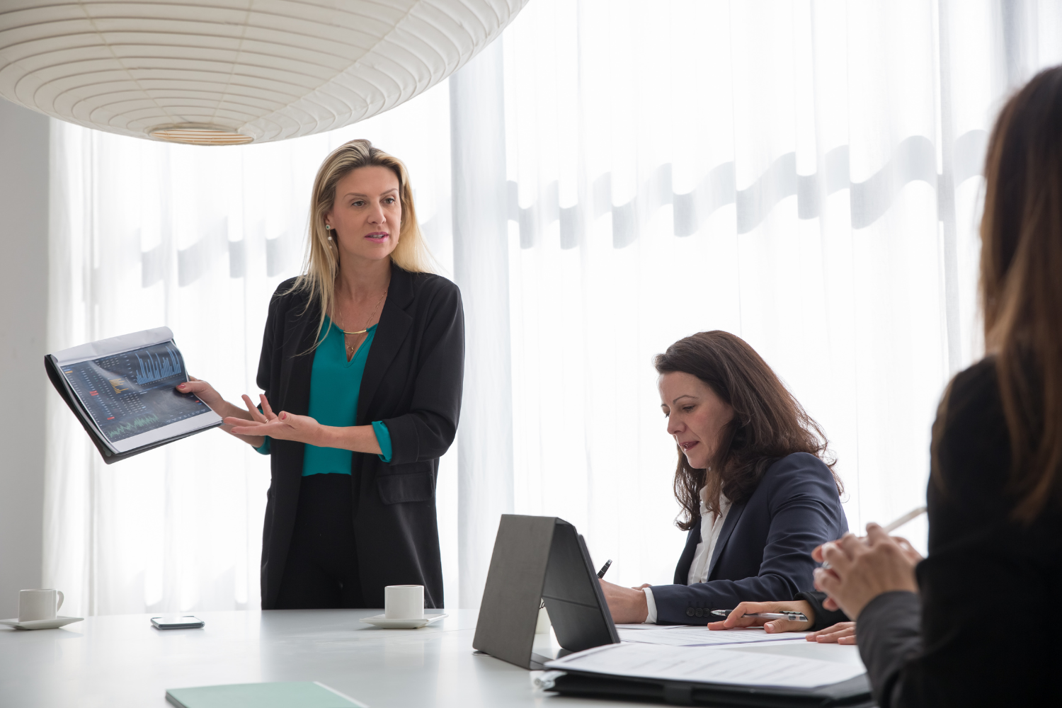 Modern office, businesswoman presenting data, colleagues seated at table, daylight-bright, documentary style, three people.