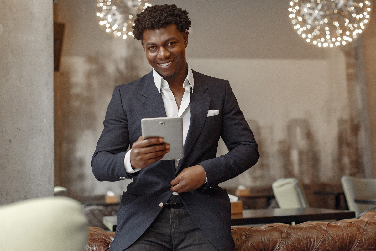 Modern café interior, man with tablet foreground, wide shot, warm ambient lighting, editorial travel photo style, single smiling figure.