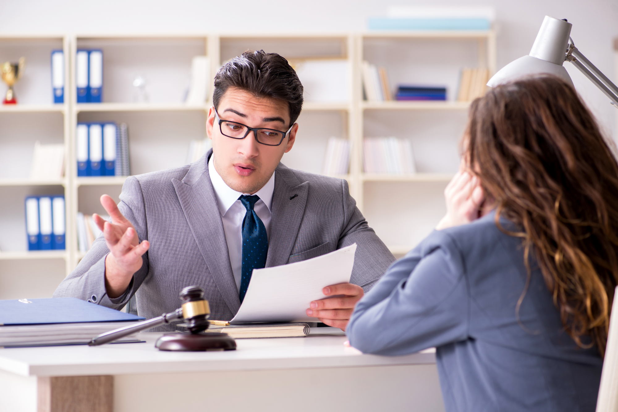 Office interior, man with papers and gavel at desk, bookshelf backdrop, daytime, documentary style, two people.