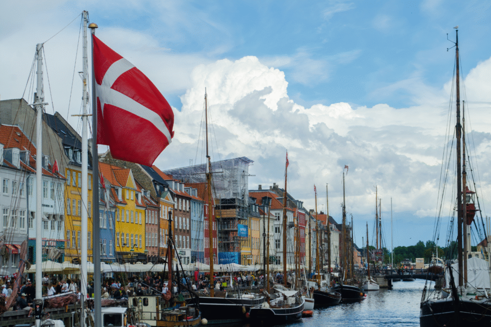 Nyhavn, boats along canal, Danish flag foreground, daytime partly cloudy, editorial travel photo, no people.