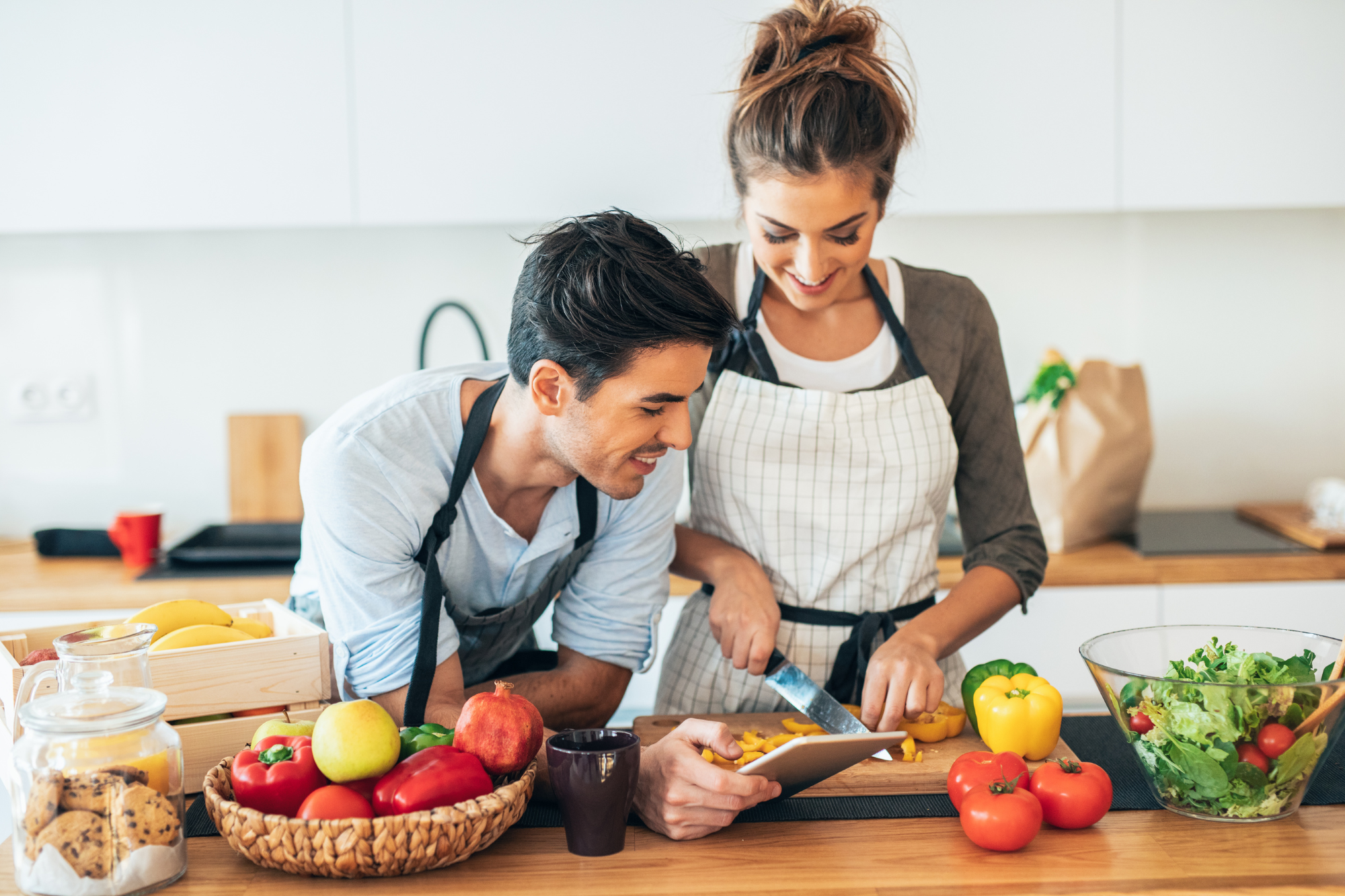 Bright kitchen, couple chopping vegetables, counter filled with produce, daytime natural light, editorial travel photo, people.