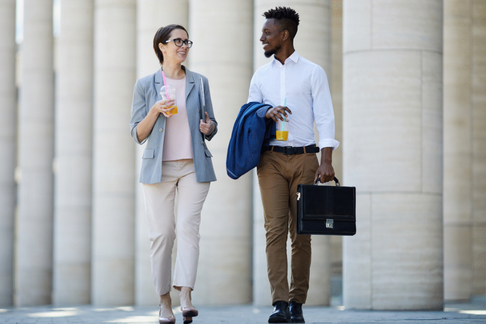 Outside office building, two professionals chatting, daytime sun, candid editorial travel photo, people smiling.