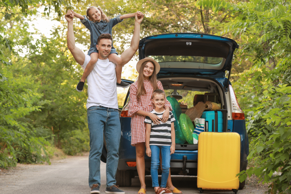Suburban driveway, family and packed car, open trunk foreground, sunny afternoon light, editorial travel photo, four people.