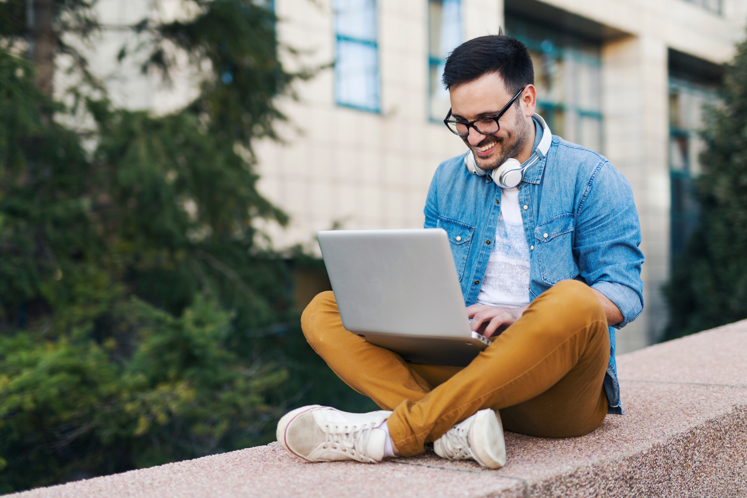 Outdoor ledge, man with glasses and headphones, cross-legged using laptop, daytime, editorial travel photo, one person.