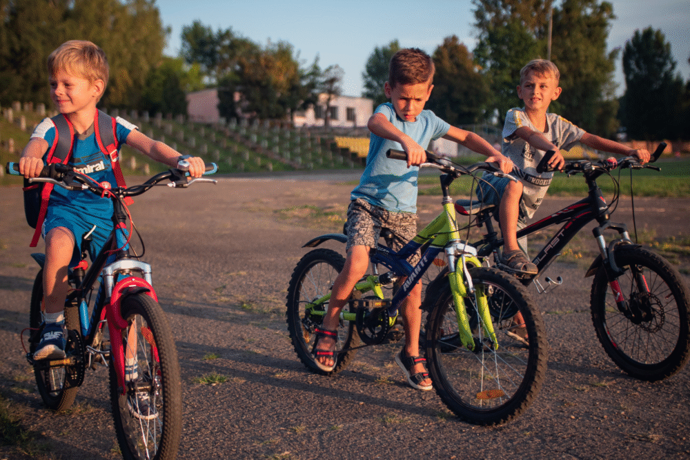 Open paved lot, three boys cycling, wide shot with background trees and building, sunny afternoon light, editorial travel photo, people.