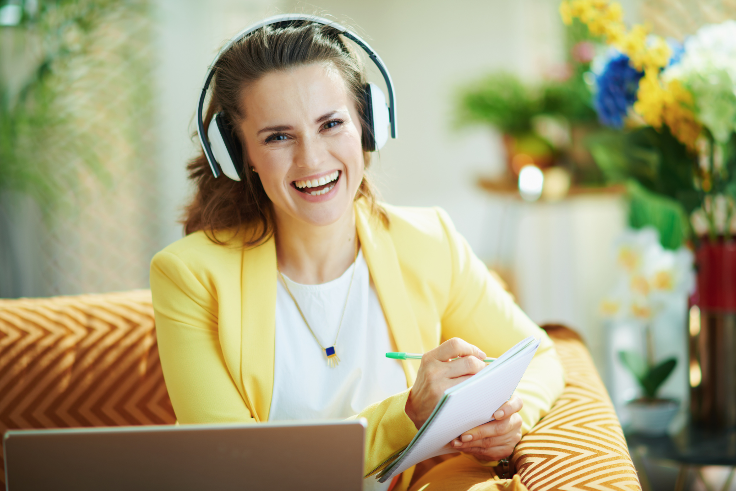 Living room, woman in yellow blazer with headphones, notepad and laptop foreground, bright daytime, editorial style, one person.
