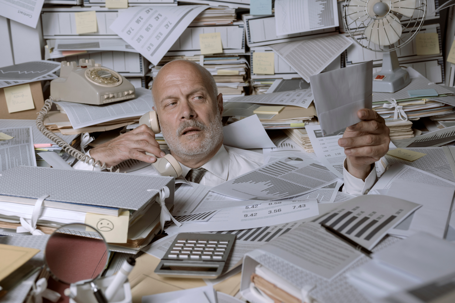 Office, overwhelmed man under paperwork mountain, cluttered desk foreground, daytime light, documentary style, people present.