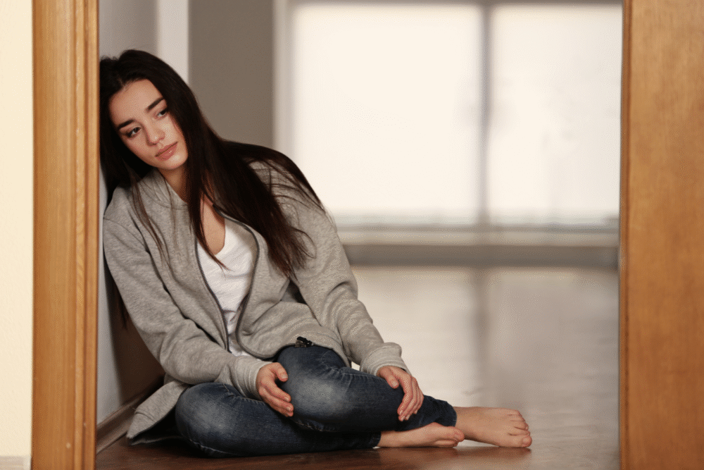 Softly lit hallway, young woman in gray hoodie and jeans, seated barefoot against wall, pensive mood, documentary style, people.