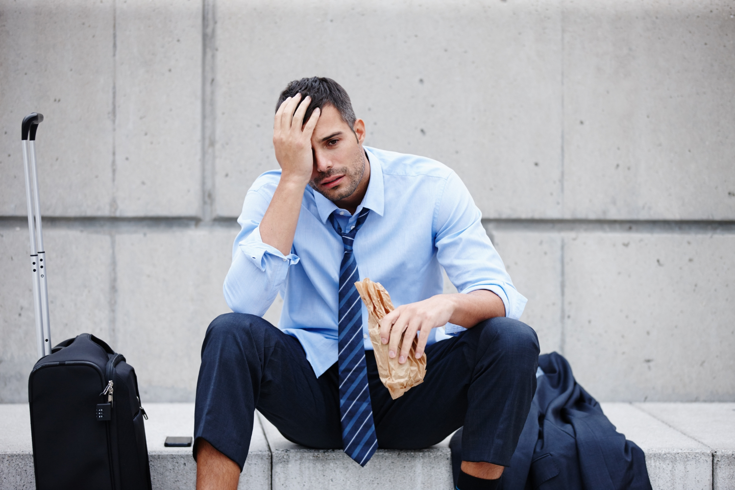 Urban plaza, stressed businessman on bench, suitcase and jacket at side, overcast daylight, editorial travel photo, single person.