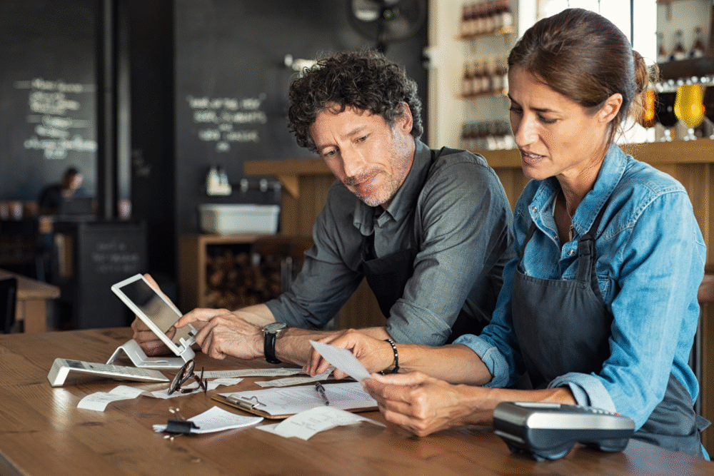 Cafe interior, receipts and tablet on table, two people in focus, daytime natural light, documentary style business scene, with people.