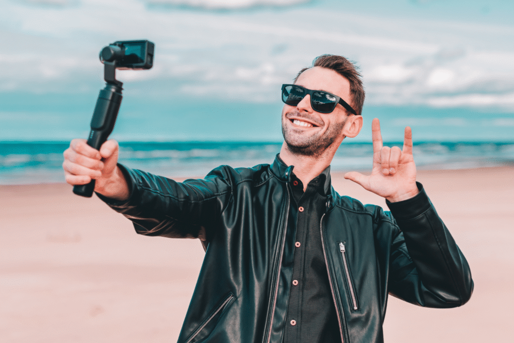Beach, man in black leather jacket and sunglasses, camera selfie with raised I love you hand, cloudy day, editorial travel photo, single person.