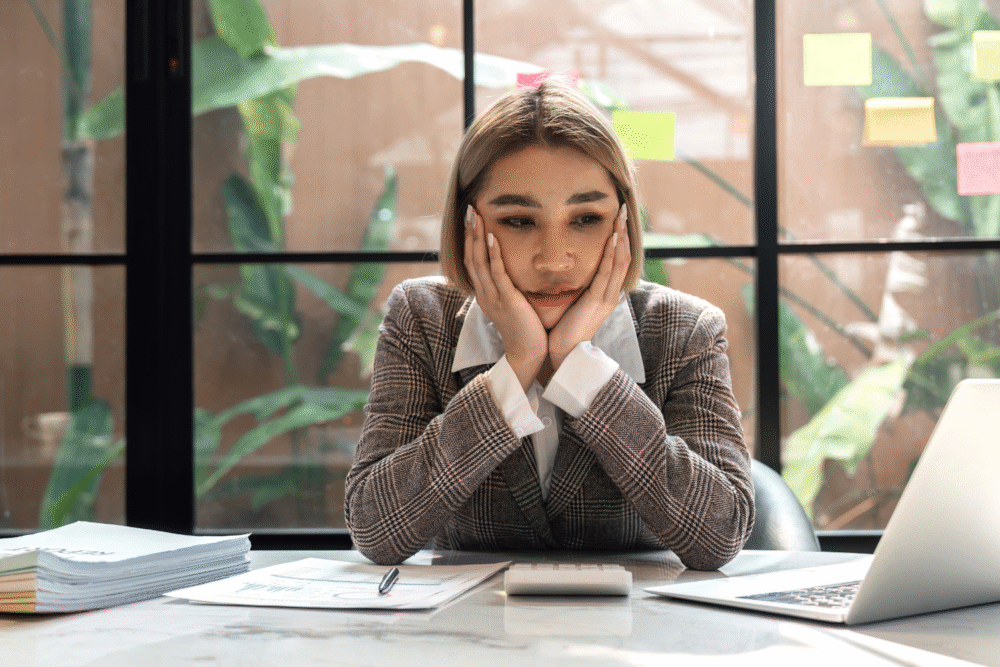 Office, woman in plaid suit at desk, chin on hands, scattered papers and tech, daytime ambient light, documentary style, one person.