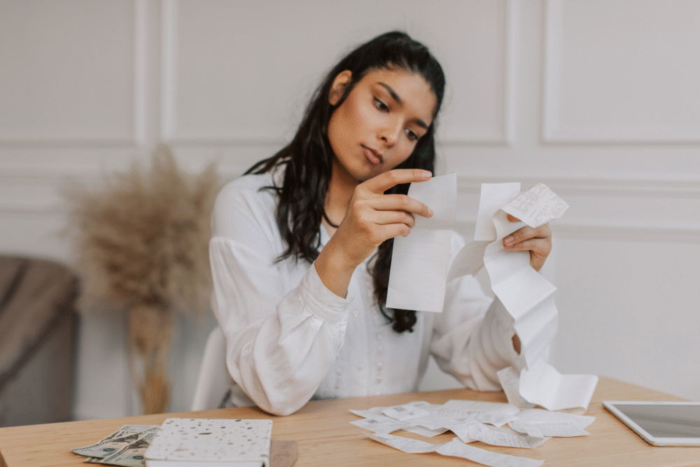 Kitchen table, woman with receipts, cluttered workspace, midday natural light, documentary style, one adult.