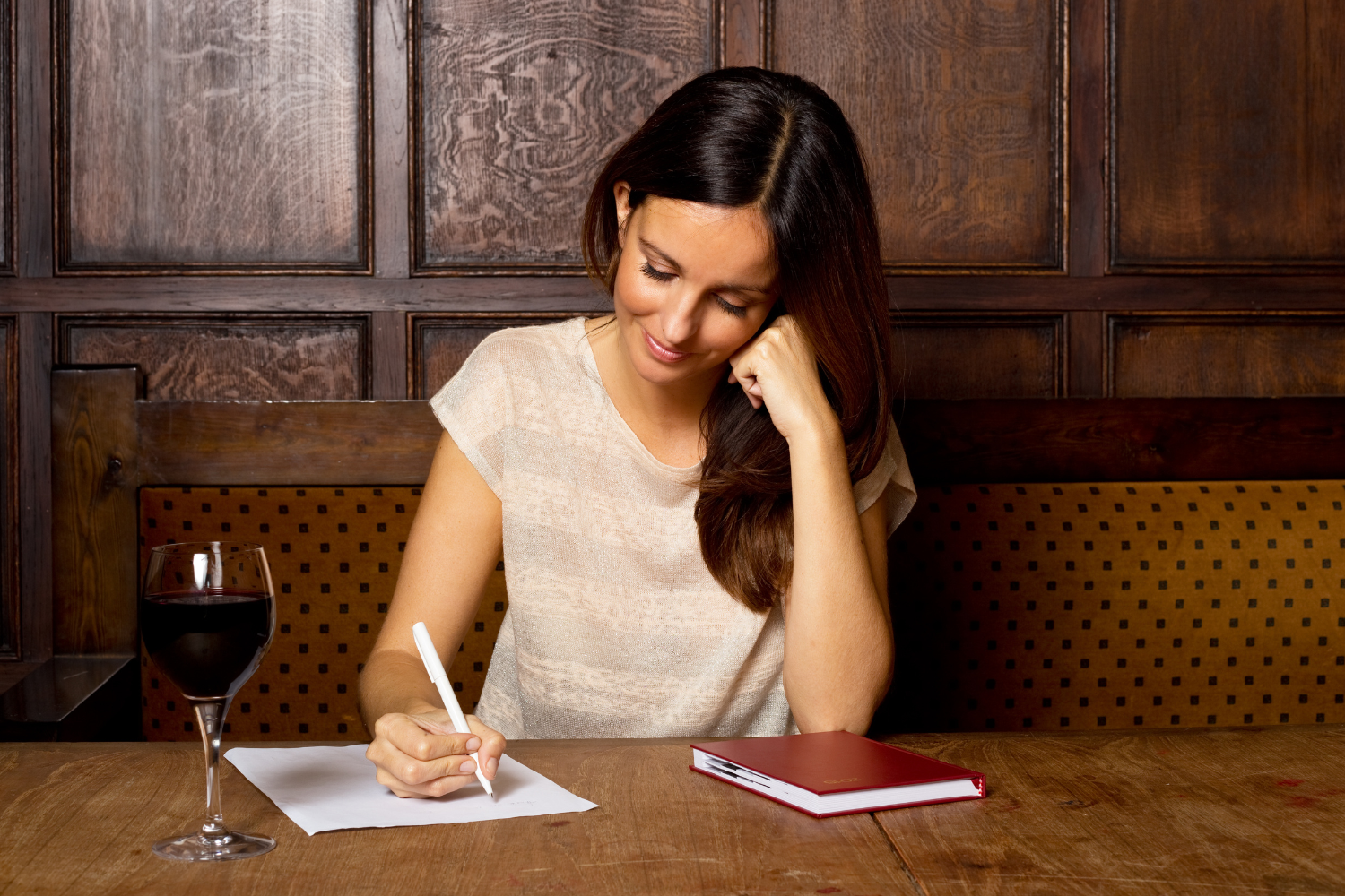 Wood-paneled room, woman writing at table, wine and notebook nearby, soft evening light, editorial travel photo, one person.