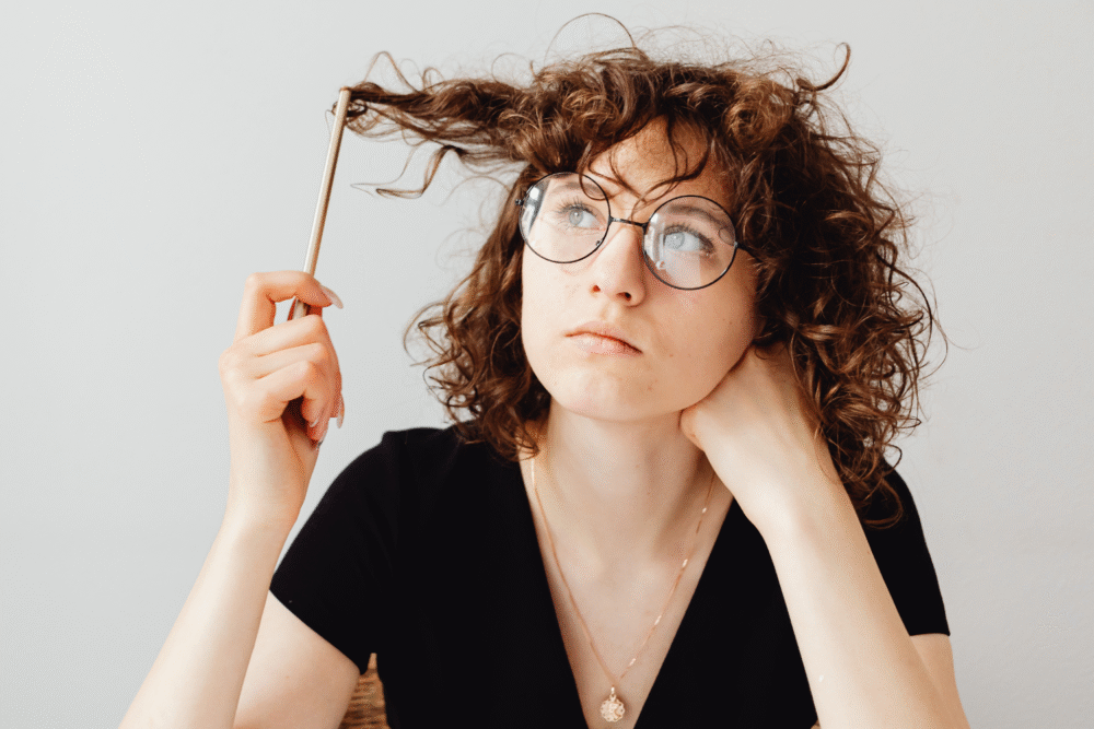 Table, young woman with curly hair and glasses, mid-shot, bright interior lighting, documentary style, person present.