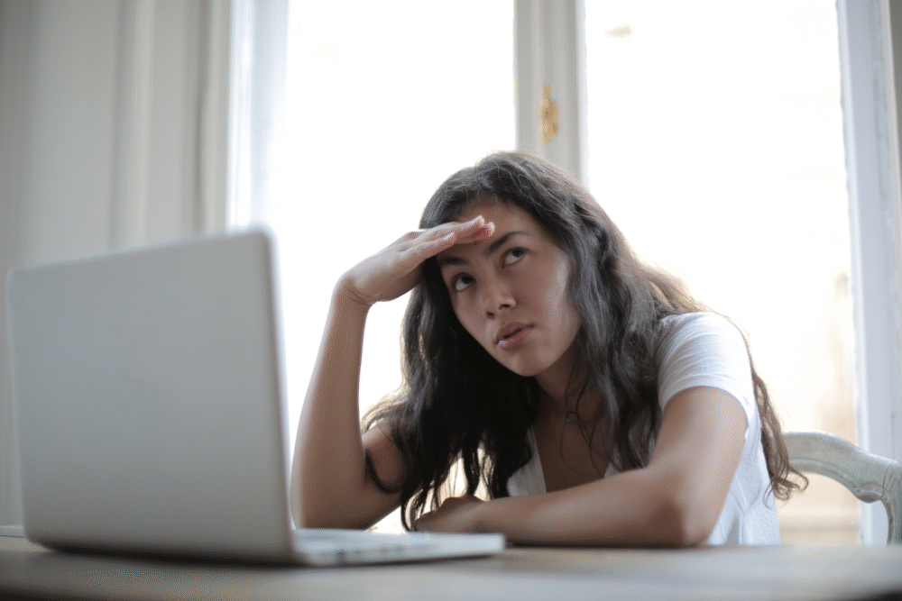 Office interior, woman shielding eyes, laptop on desk foreground, daylight through window, documentary style, single person.