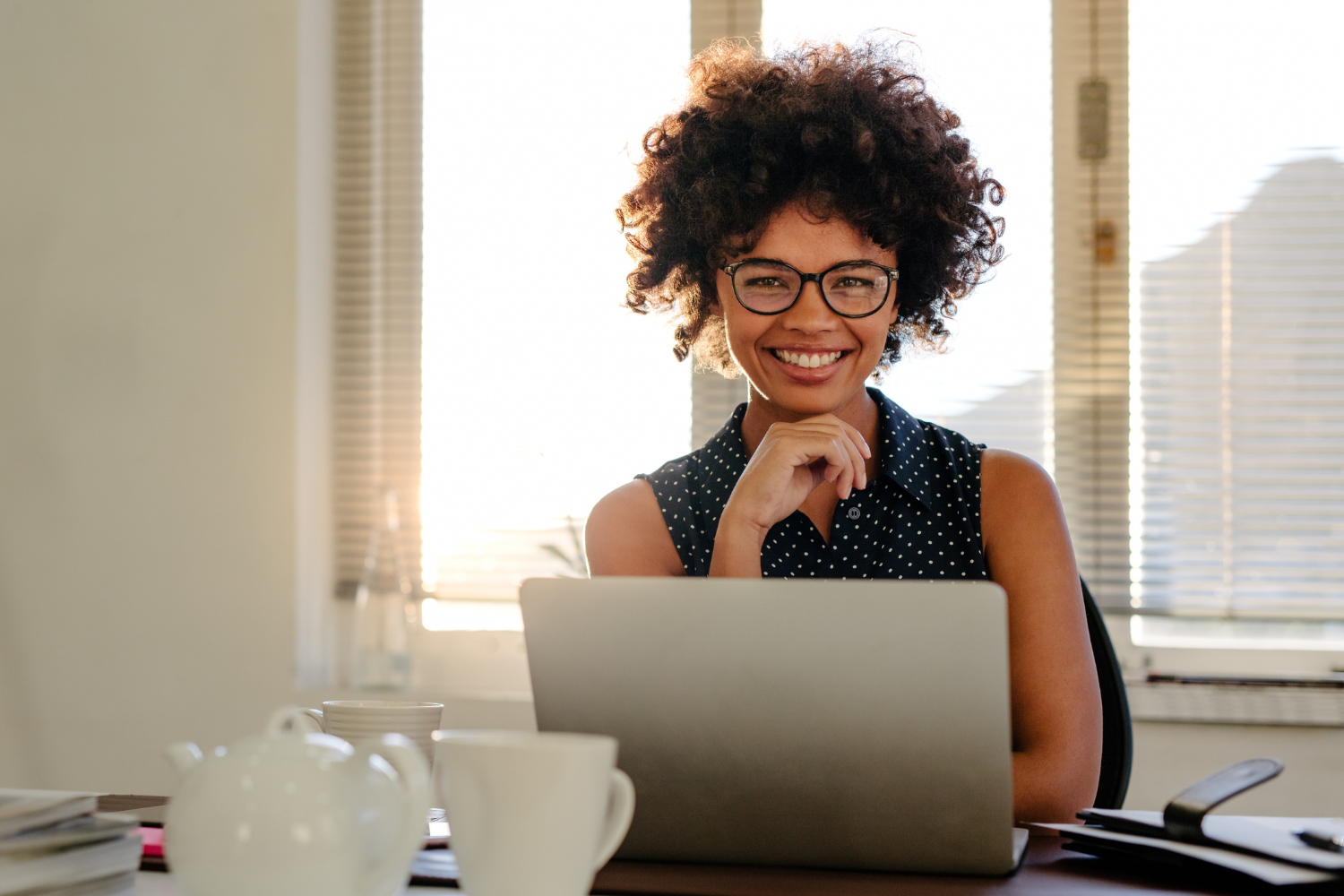 Desk workspace, woman with curly hair and glasses, laptop center frame, morning sunlight streaming, documentary style, one person.